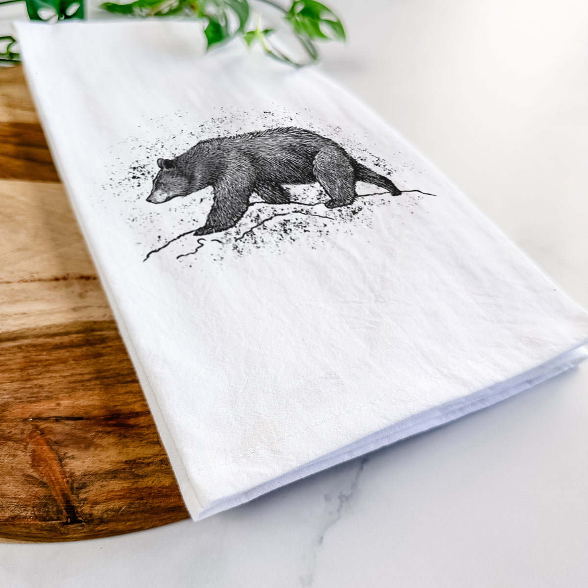 Close-up of a folded white tea towel on a wooden board, showing a detailed black ink sketch of a black bear walking across rocky ground.