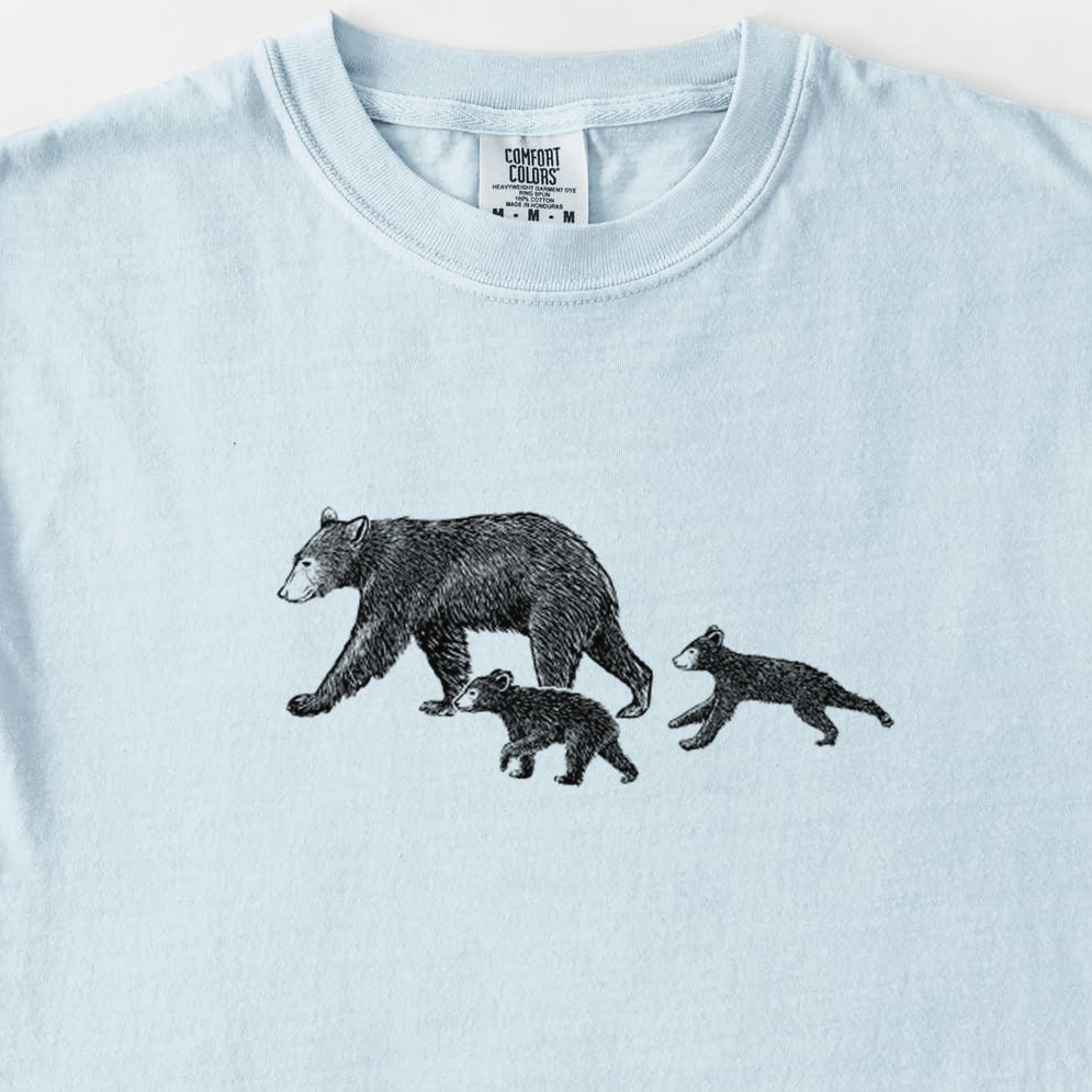 Chambray blue Comfort Colors t-shirt laid flat, printed with a black ink illustration of a mother black bear walking with two cubs.