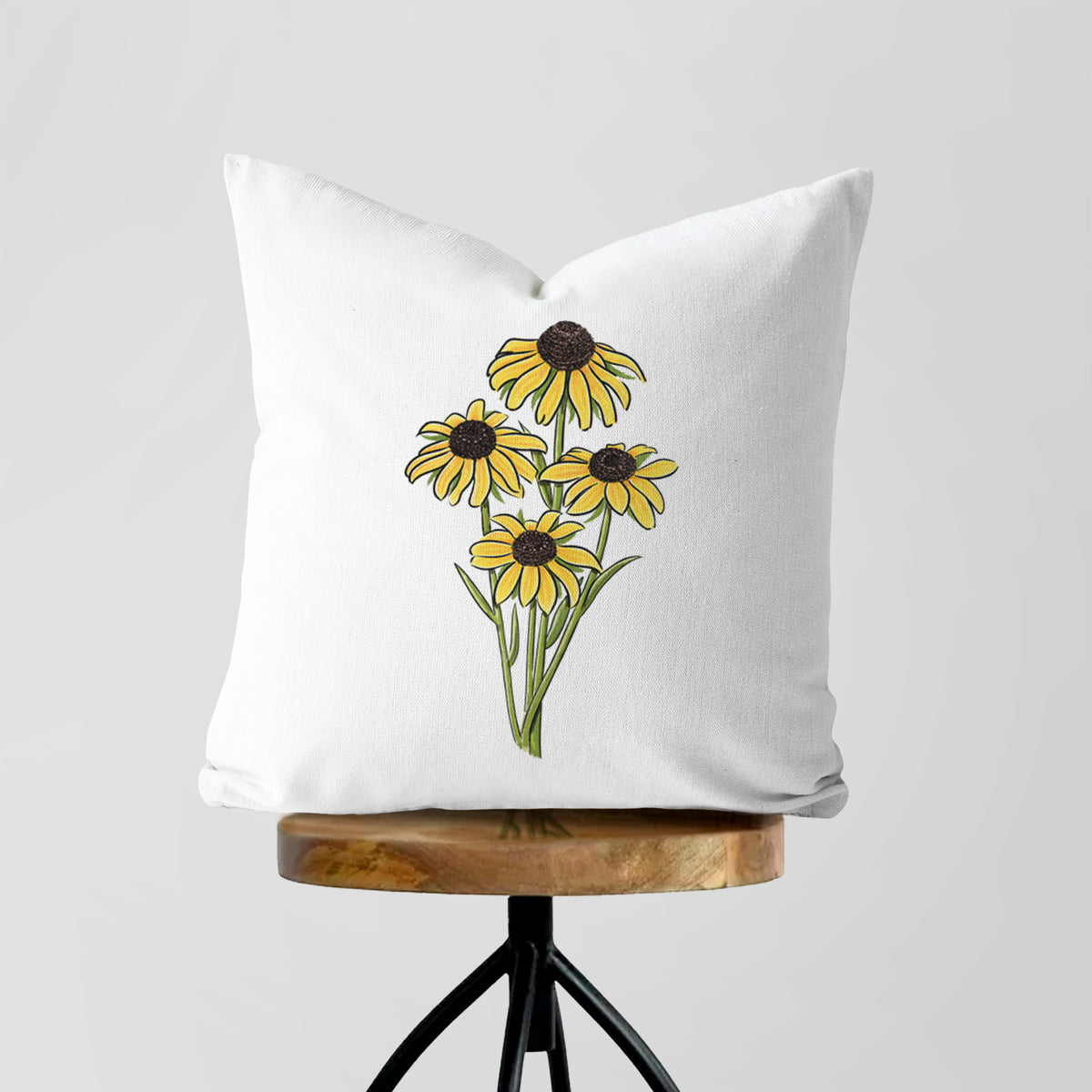White pillow cover printed with a full-color bouquet of yellow black-eyed Susan flowers, on a wooden stool.