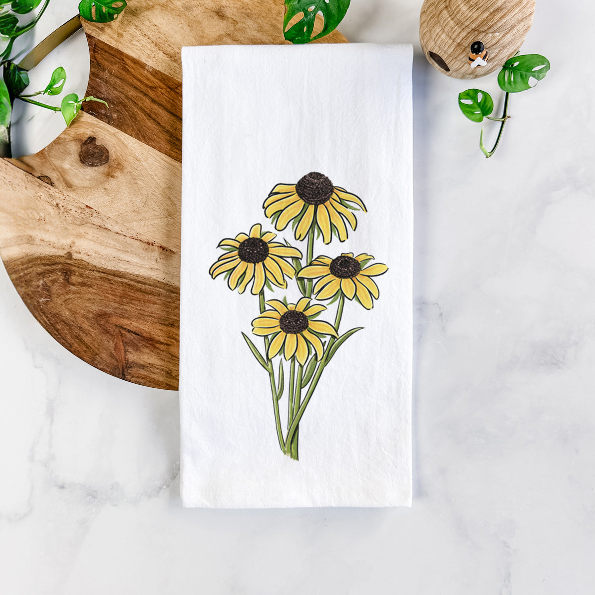 White cotton tea towel printed with a full-color bouquet of yellow black-eyed Susan flowers, on a marble counter beside a round wooden cutting board and greenery.