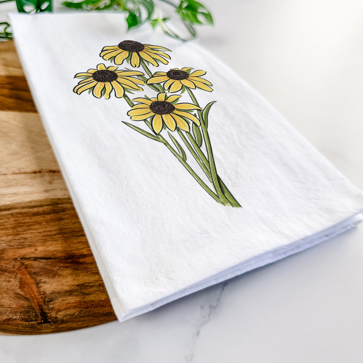 Close-up of a folded white tea towel on a wooden board, showing a full-color bouquet of yellow black-eyed Susan flowers.