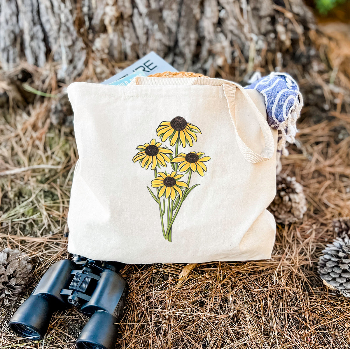 Canvas tote bag printed with a full-color bouquet of yellow black-eyed Susan flowers, resting on dry grass beside a pair of binoculars.
