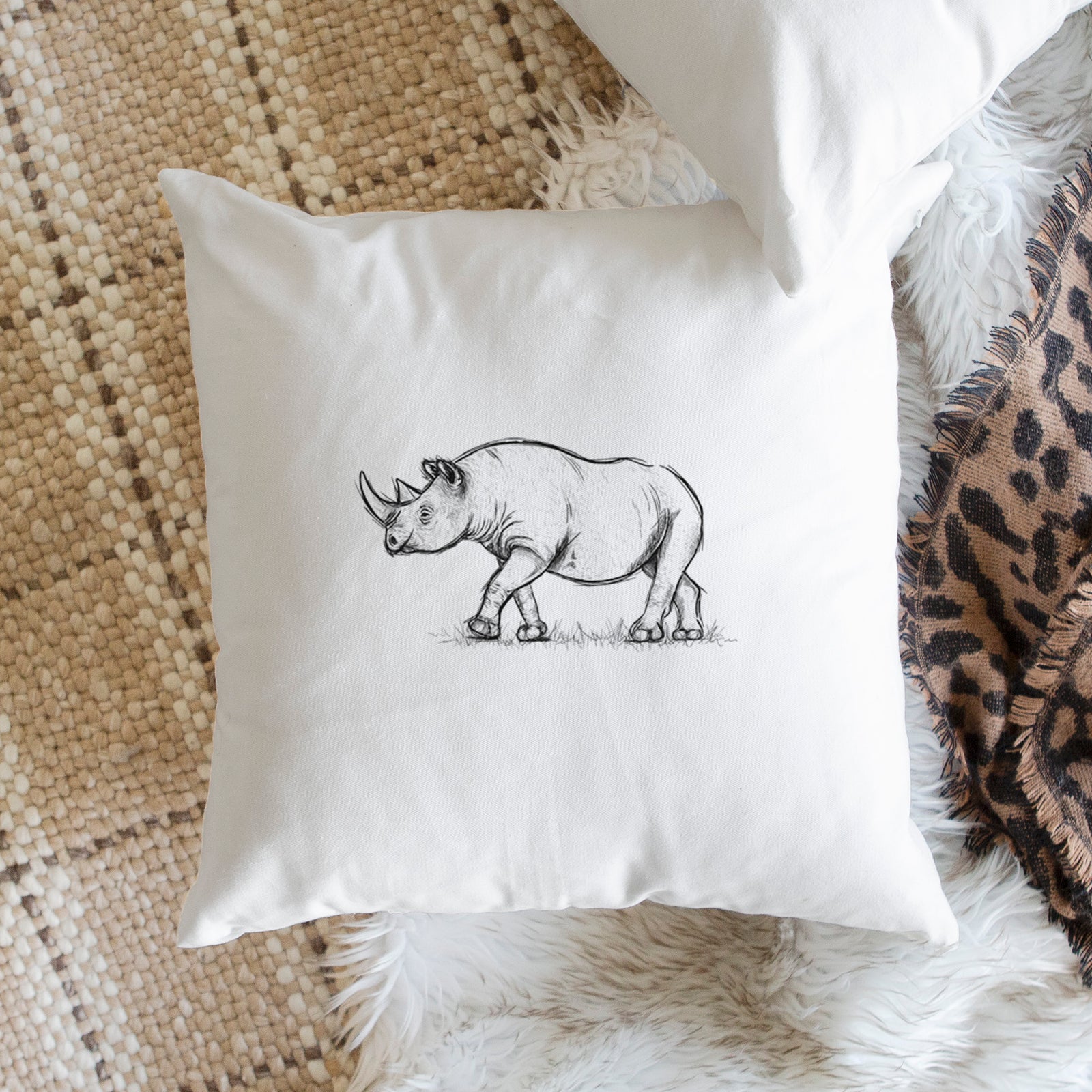 White pillow cover printed with a black ink illustration of a black rhinoceros walking, on a bench beside plants.