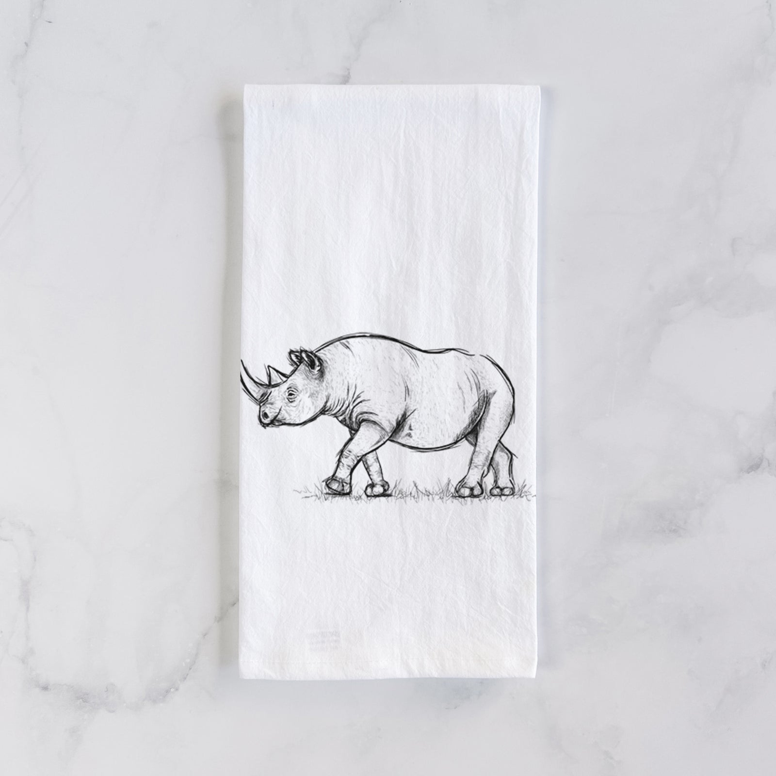 White cotton tea towel printed with a black ink illustration of a black rhinoceros walking, on a marble counter beside a round wooden cutting board and greenery.