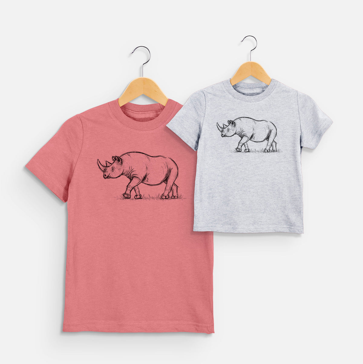 Two youth t-shirts in different colors on hangers, printed with a black ink illustration of a black rhinoceros walking.