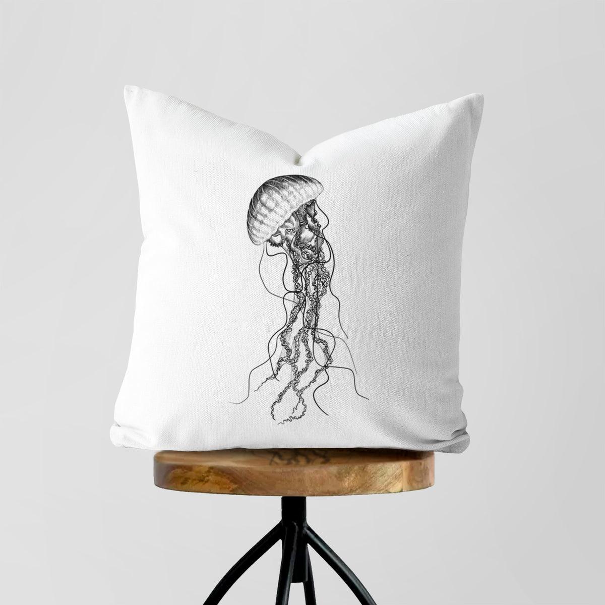 White pillow cover printed with a black ink illustration of a black sea nettle jellyfish with long trailing tentacles, on a wooden stool.