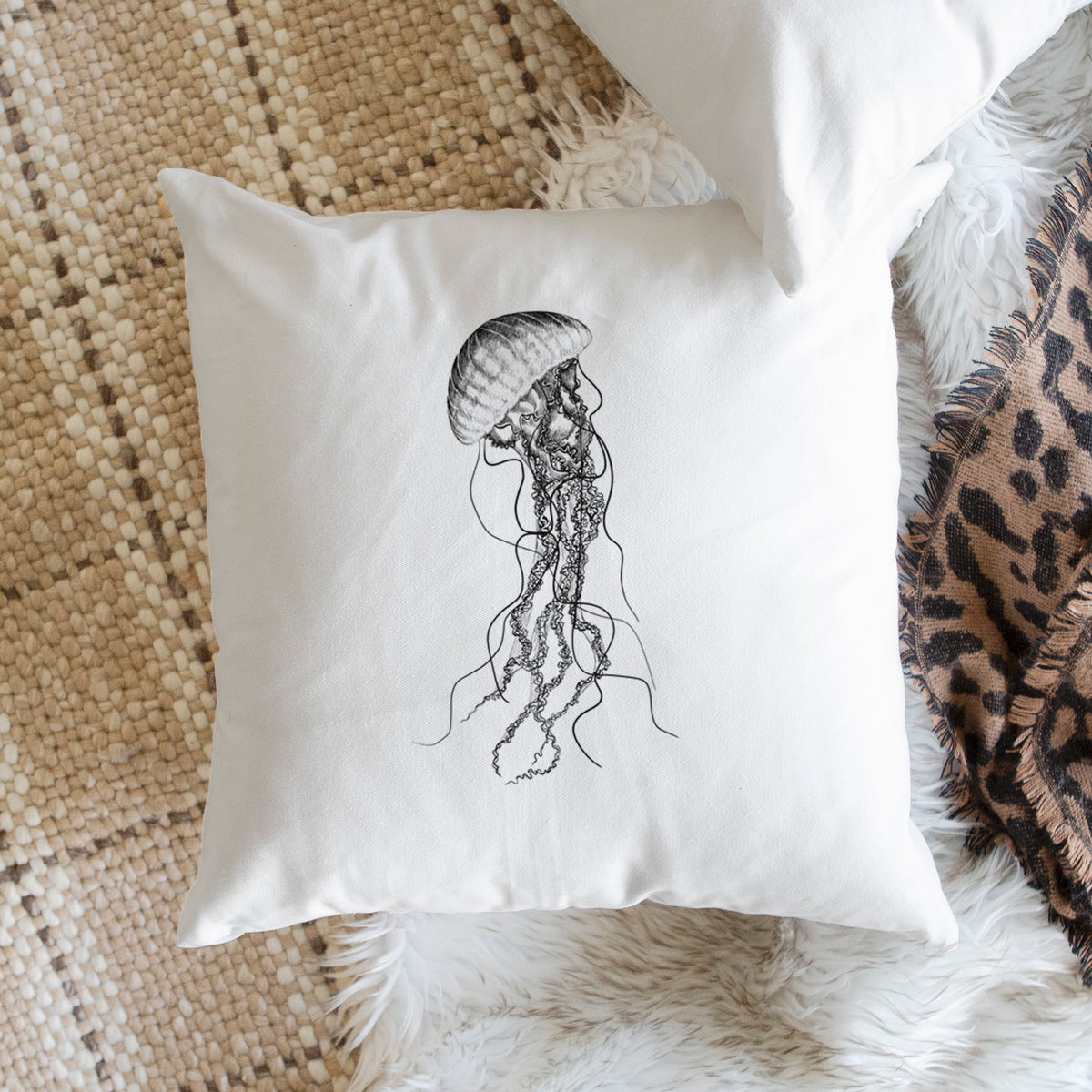 White pillow cover printed with a black ink illustration of a black sea nettle jellyfish with long trailing tentacles, on a woven chair.