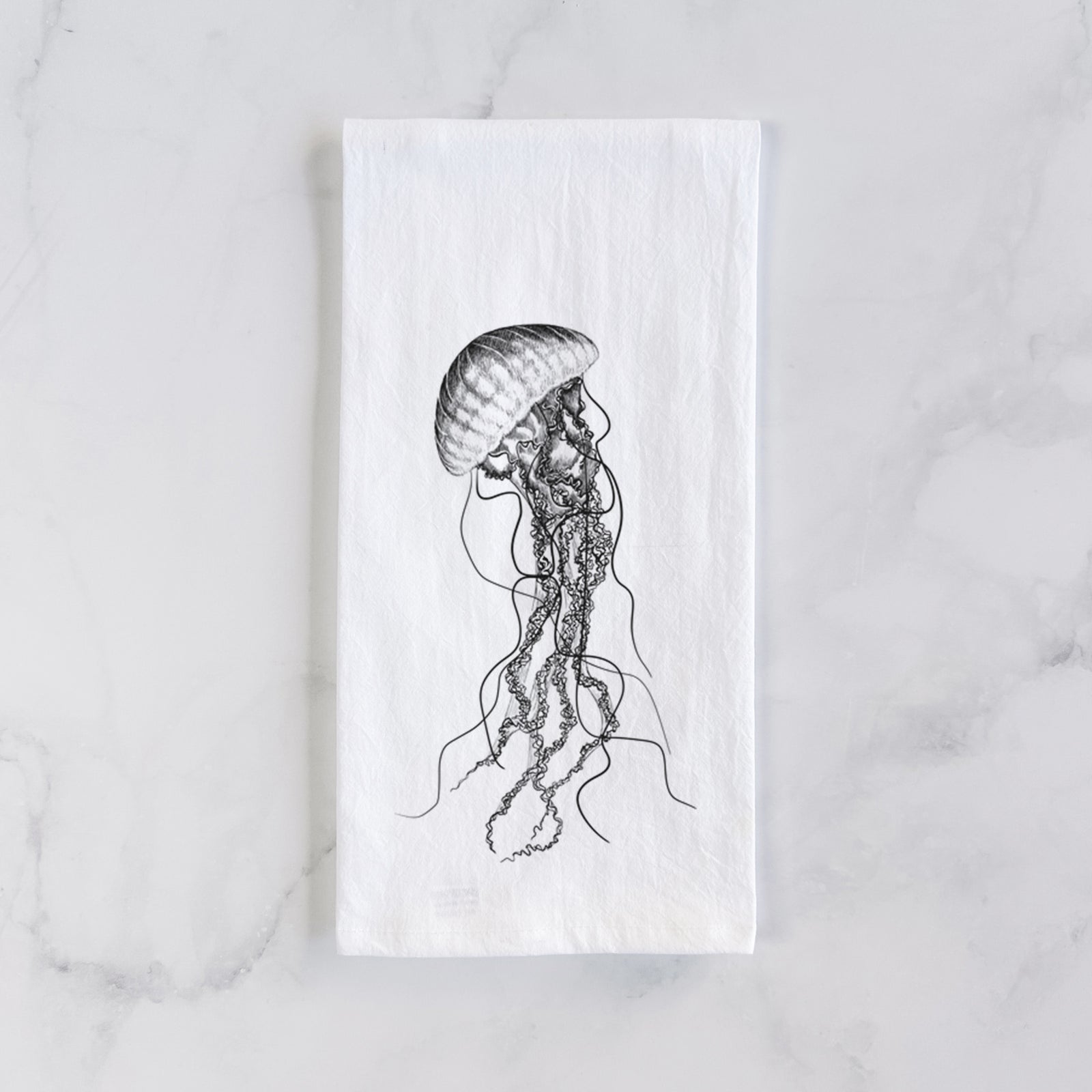 White cotton tea towel printed with a black ink illustration of a black sea nettle jellyfish with long trailing tentacles, on a marble counter beside a round wooden cutting board and greenery.