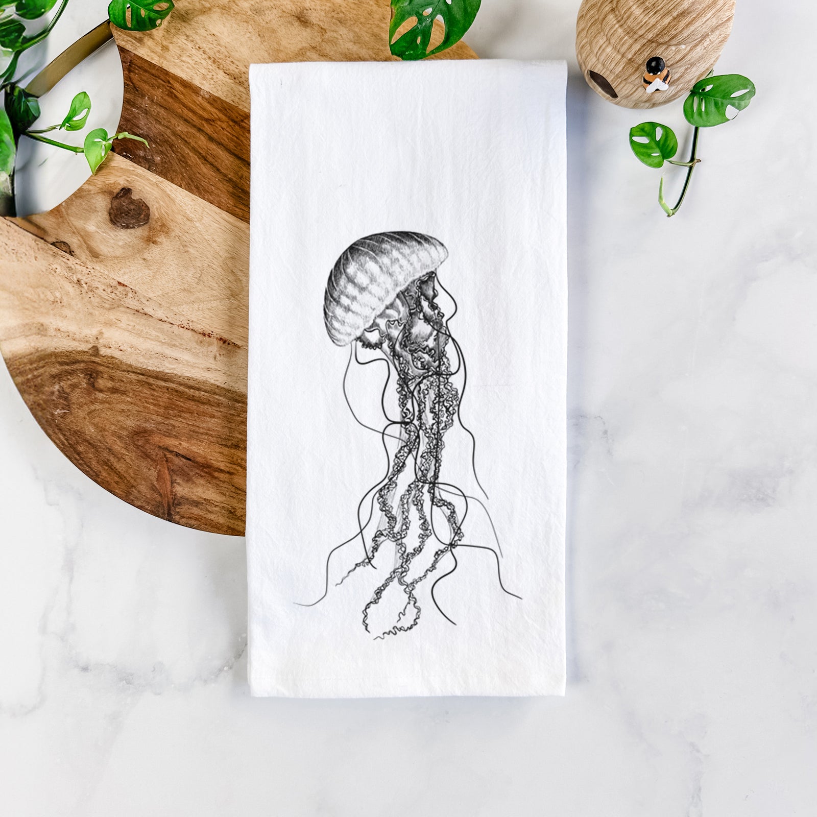 White cotton tea towel printed with a black ink illustration of a black sea nettle jellyfish with long trailing tentacles, on a marble counter beside a round wooden cutting board and greenery.