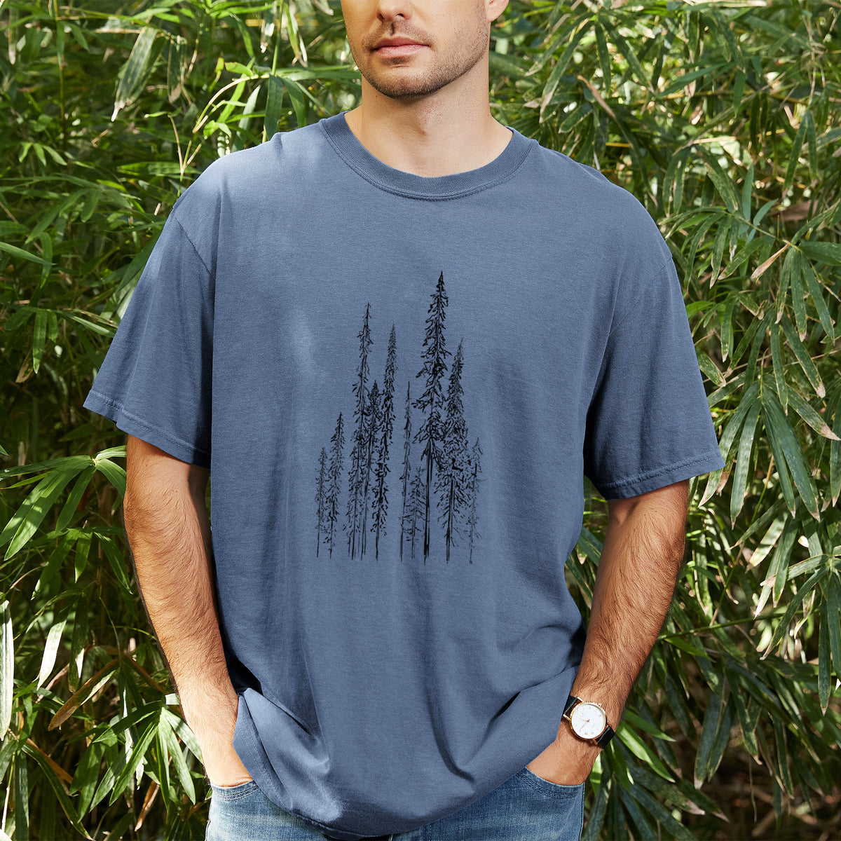 Man wearing a Comfort Colors t-shirt printed with a black ink illustration of a stand of tall, narrow black spruce trees, outdoors.