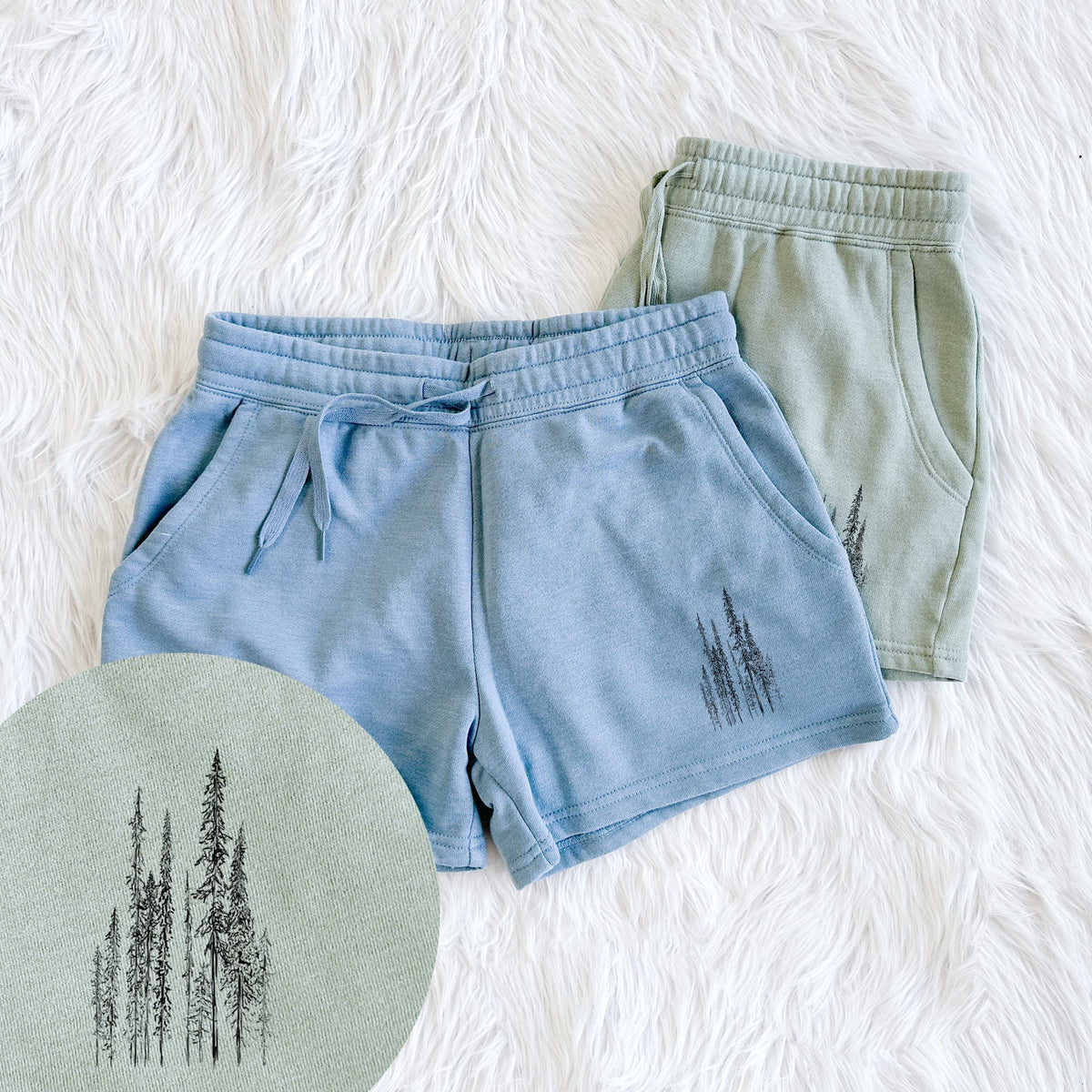 Two pairs of Cali Wave shorts in different colors printed with a black ink illustration of a stand of tall, narrow black spruce trees, with a close-up of the print.