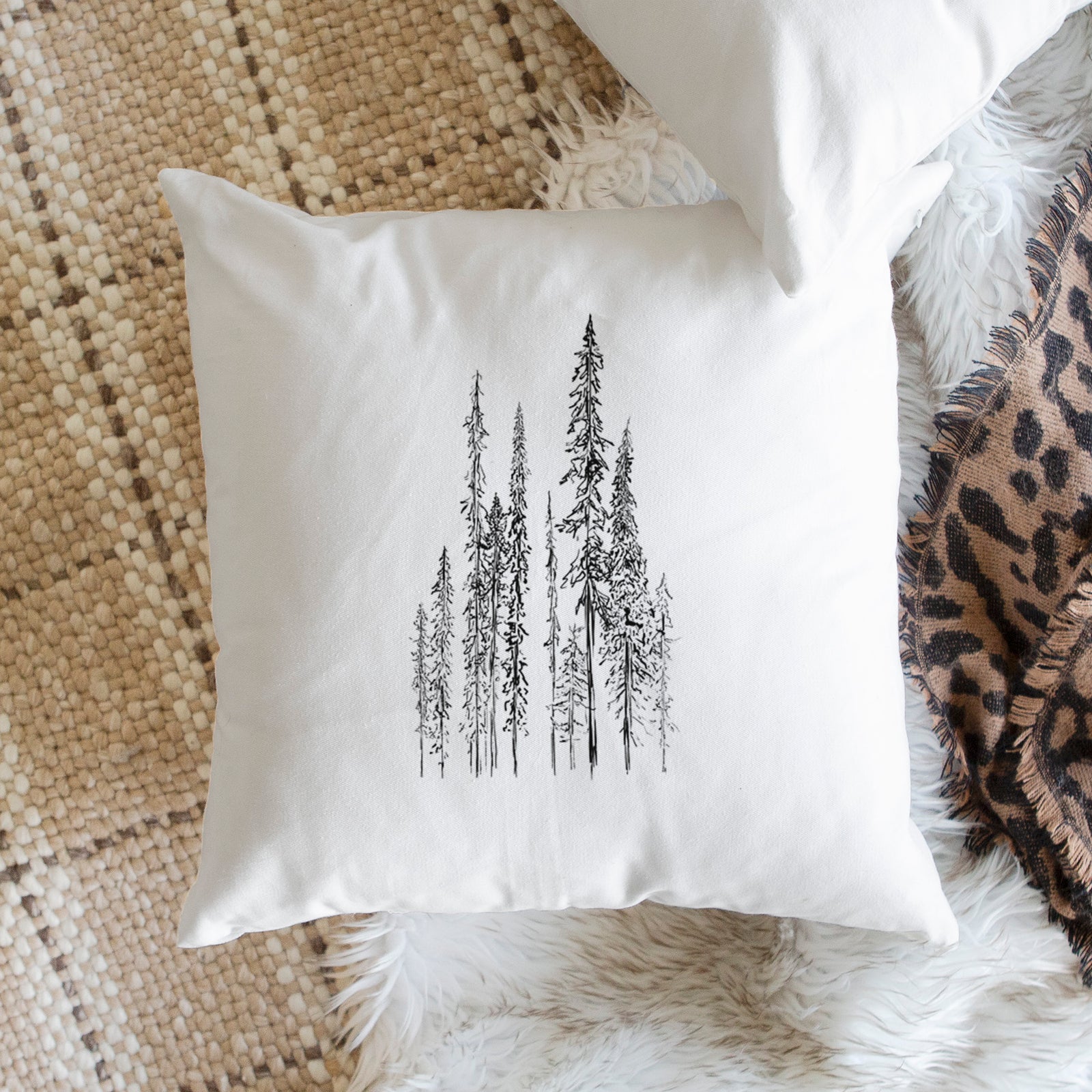 White pillow cover printed with a black ink illustration of a stand of tall, narrow black spruce trees, on a bench beside plants.