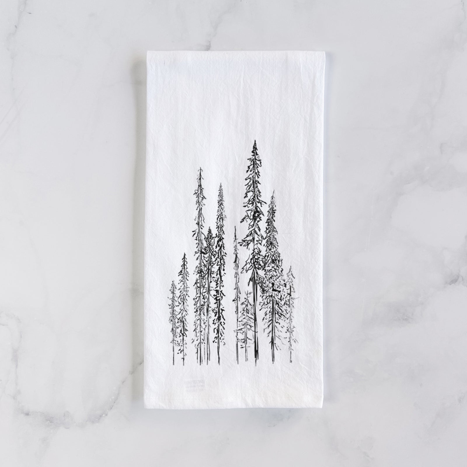 White cotton tea towel printed with a black ink illustration of a stand of tall, narrow black spruce trees, on a marble counter beside a round wooden cutting board and greenery.