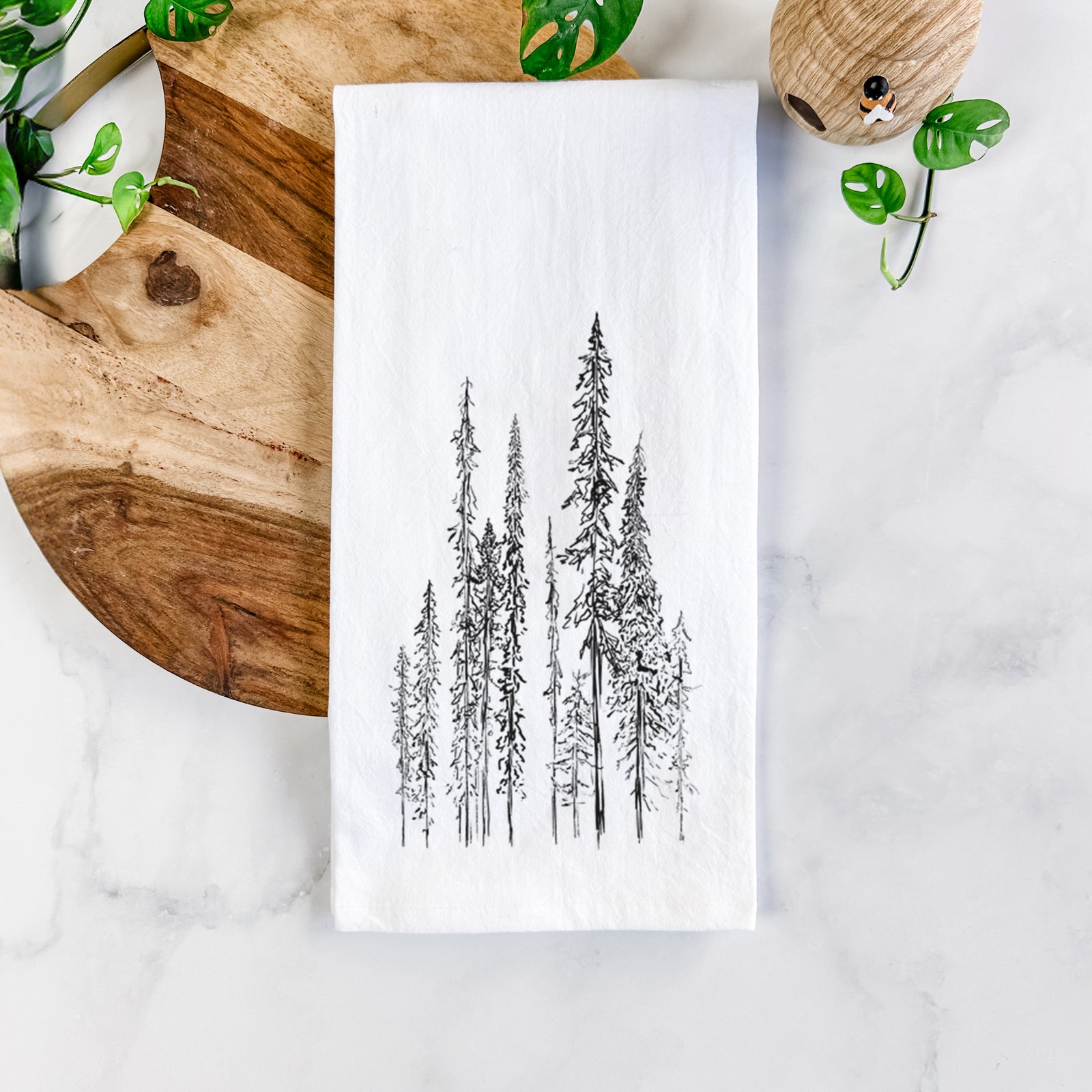 White cotton tea towel printed with a black ink illustration of a stand of tall, narrow black spruce trees, on a marble counter beside a round wooden cutting board and greenery.