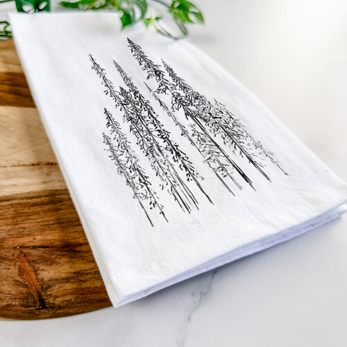 Close-up of a folded white tea towel on a wooden board, showing a black ink illustration of a stand of tall, narrow black spruce trees.