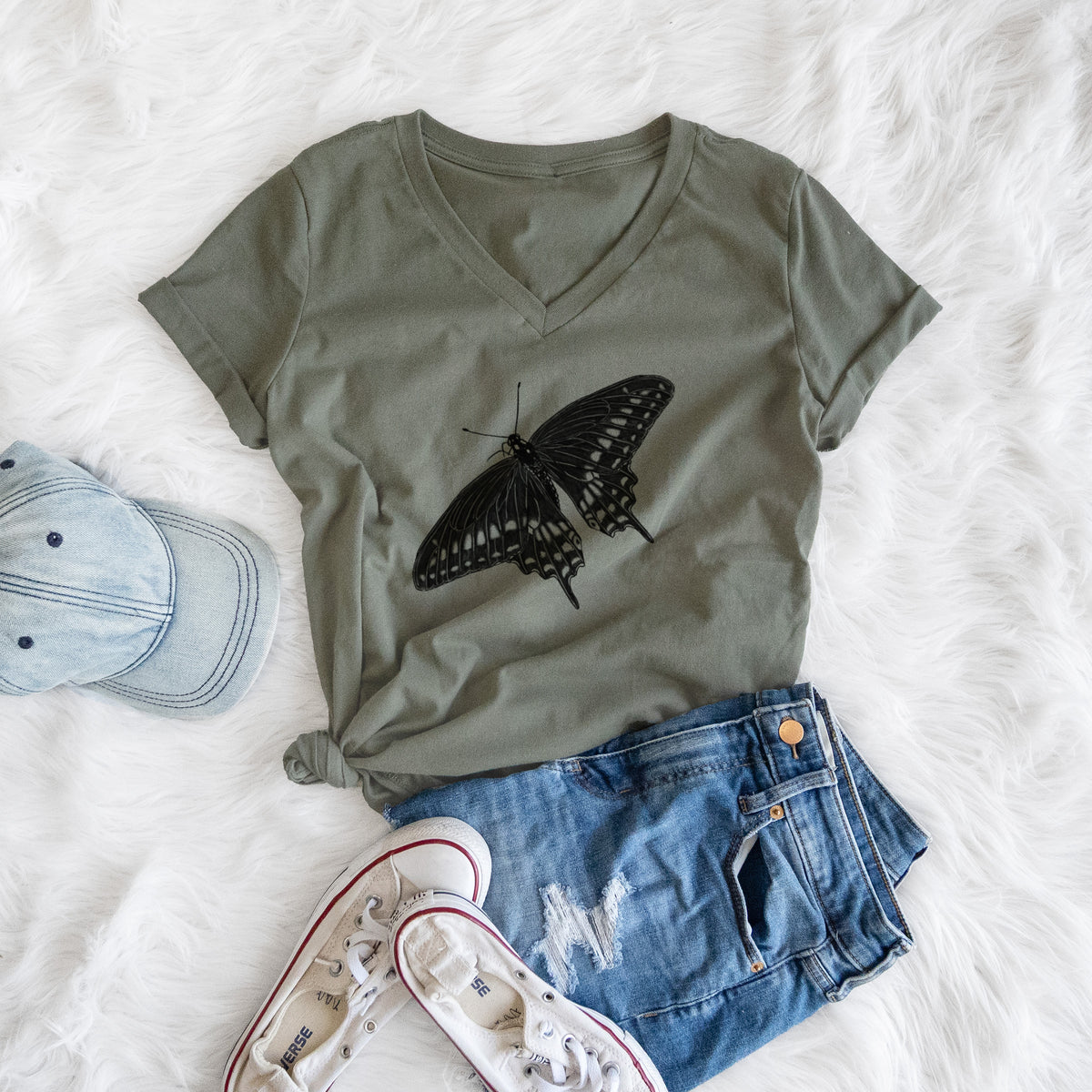 Military green women's relaxed fit cotton V-neck t-shirt printed with a detailed black ink illustration of a black swallowtail butterfly with its wings spread, styled flat on a white faux fur rug with a cap, jeans and sneakers.