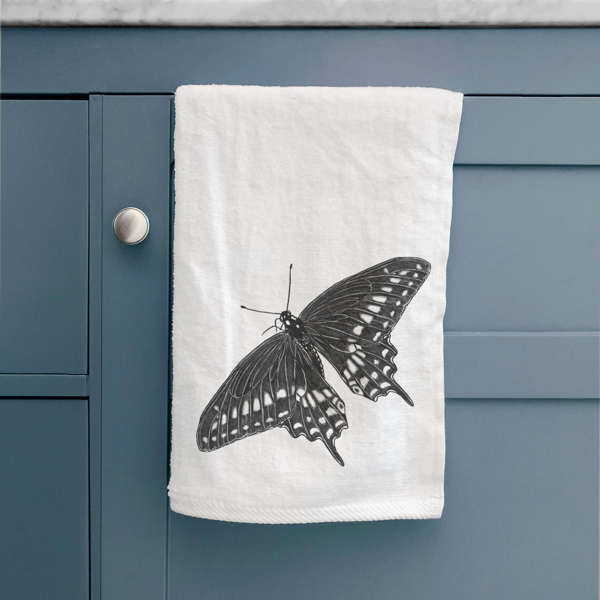 White hand towel printed with a detailed black ink illustration of a black swallowtail butterfly with its wings spread, hanging on a cabinet door.