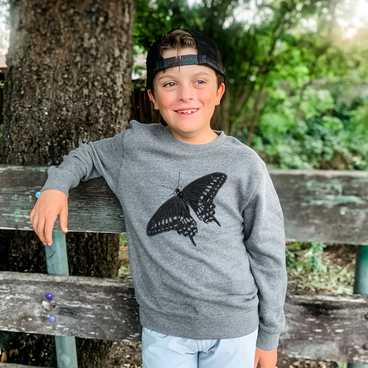 Boy wearing a youth crewneck sweatshirt printed with a detailed black ink illustration of a black swallowtail butterfly with its wings spread, outdoors.