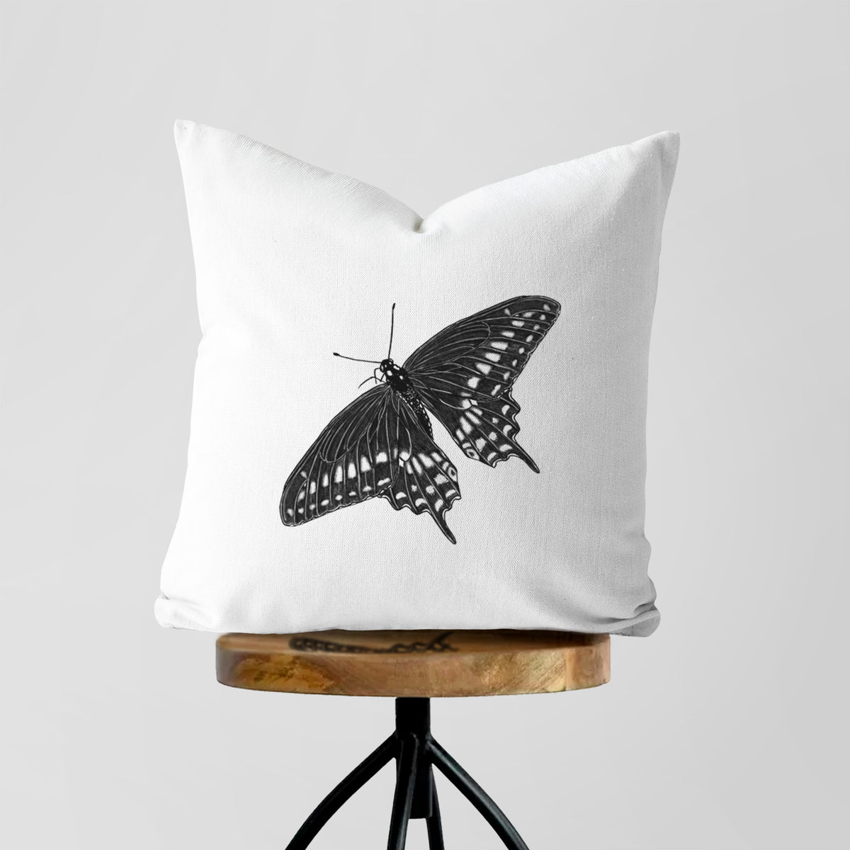 White pillow cover printed with a detailed black ink illustration of a black swallowtail butterfly with its wings spread, on a wooden stool.