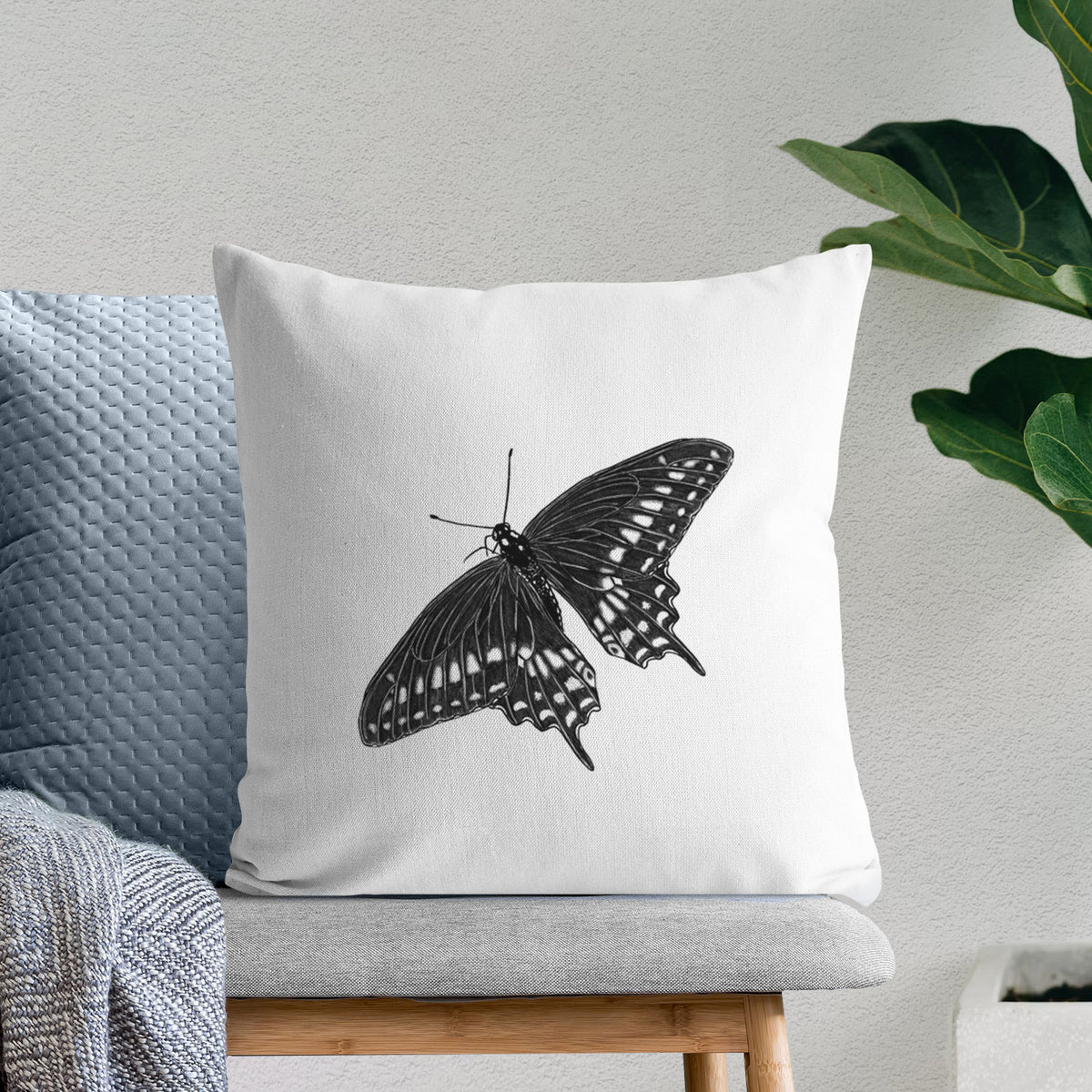 White pillow cover printed with a detailed black ink illustration of a black swallowtail butterfly with its wings spread, on a bench beside plants.