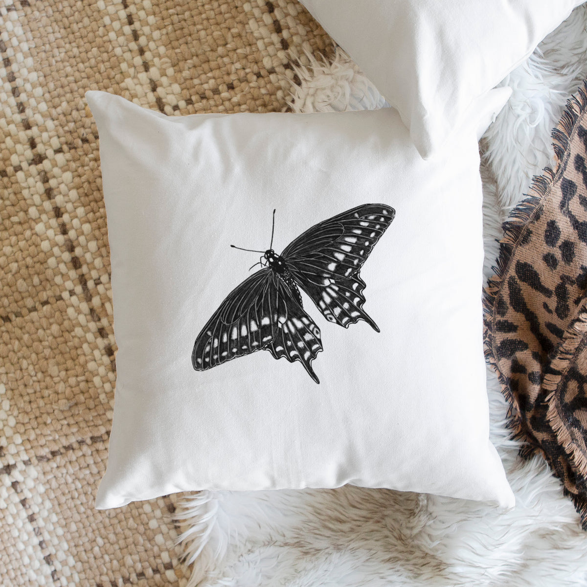 White pillow cover printed with a detailed black ink illustration of a black swallowtail butterfly with its wings spread, on a woven chair.