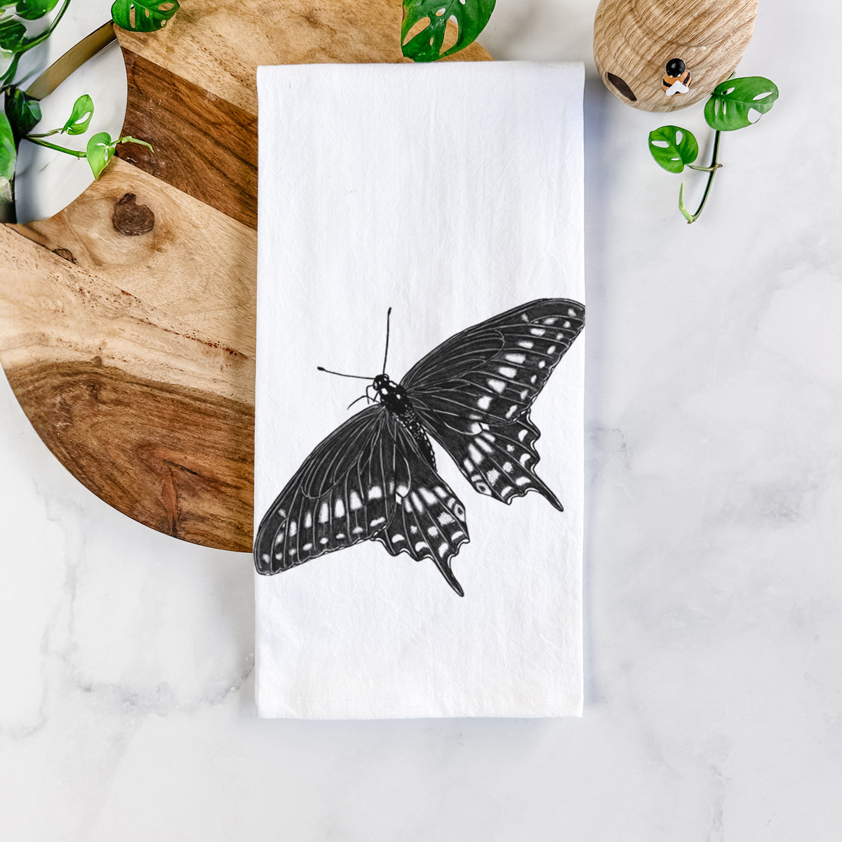 White cotton tea towel printed with a detailed black ink illustration of a black swallowtail butterfly with its wings spread, on a marble counter beside a round wooden cutting board and greenery.