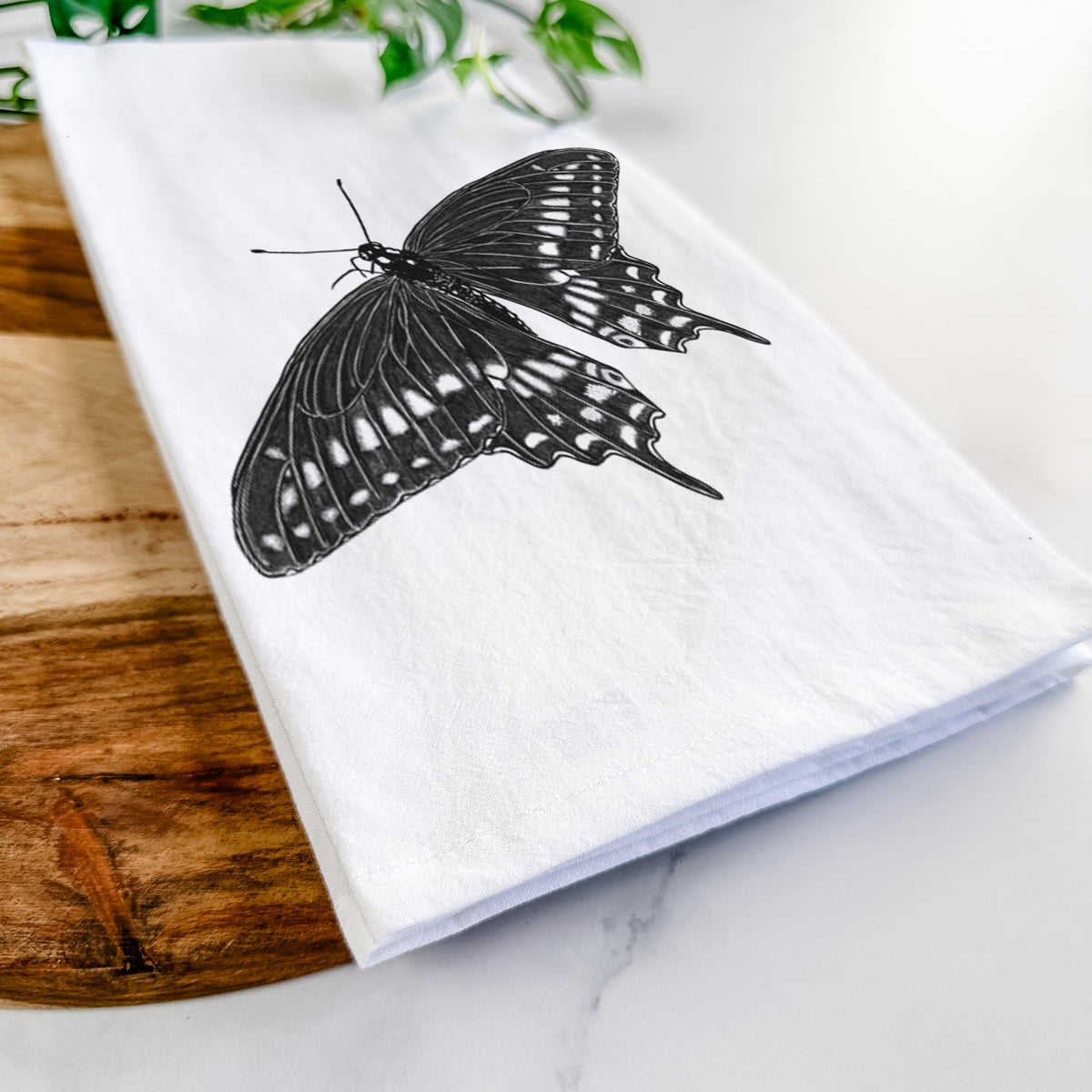 Close-up of a folded white tea towel on a wooden board, showing a detailed black ink illustration of a black swallowtail butterfly with its wings spread.
