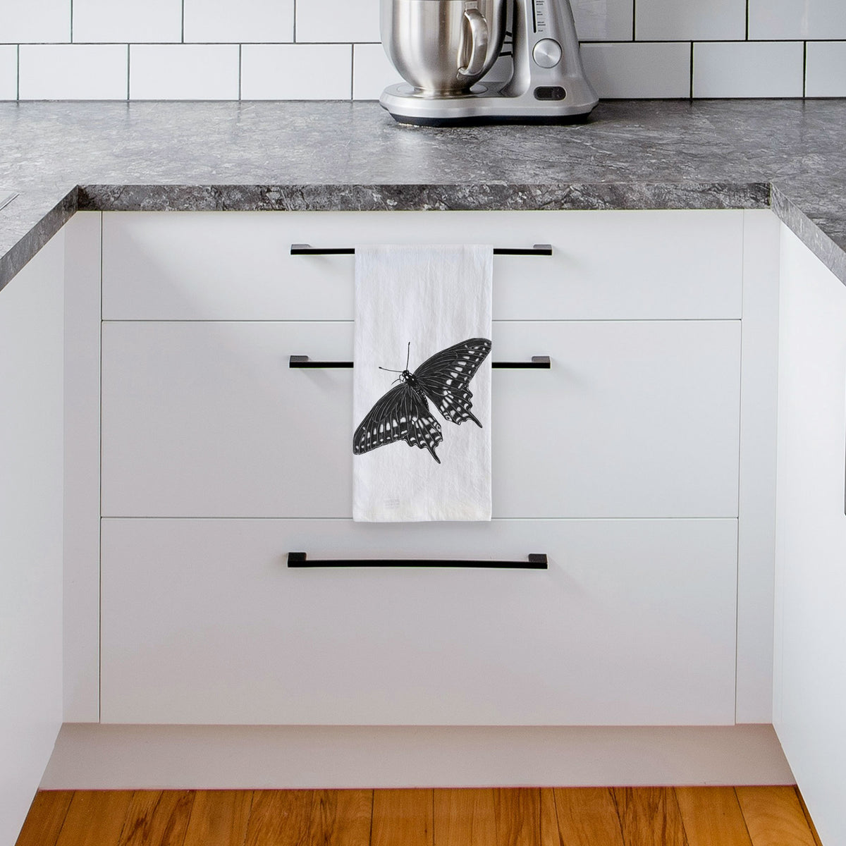 White tea towel printed with a detailed black ink illustration of a black swallowtail butterfly with its wings spread, hanging from a drawer handle in a bright white kitchen.