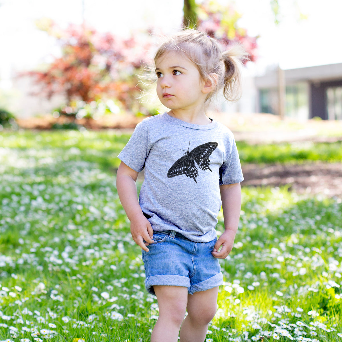 Toddler girl walking outdoors wearing a t-shirt printed with a detailed black ink illustration of a black swallowtail butterfly with its wings spread.