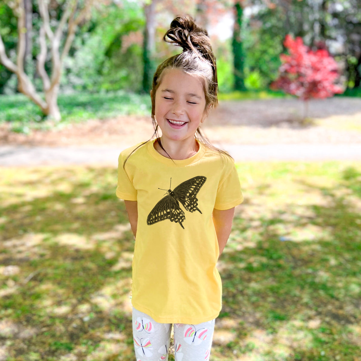 Smiling girl wearing a youth t-shirt printed with a detailed black ink illustration of a black swallowtail butterfly with its wings spread, outdoors.