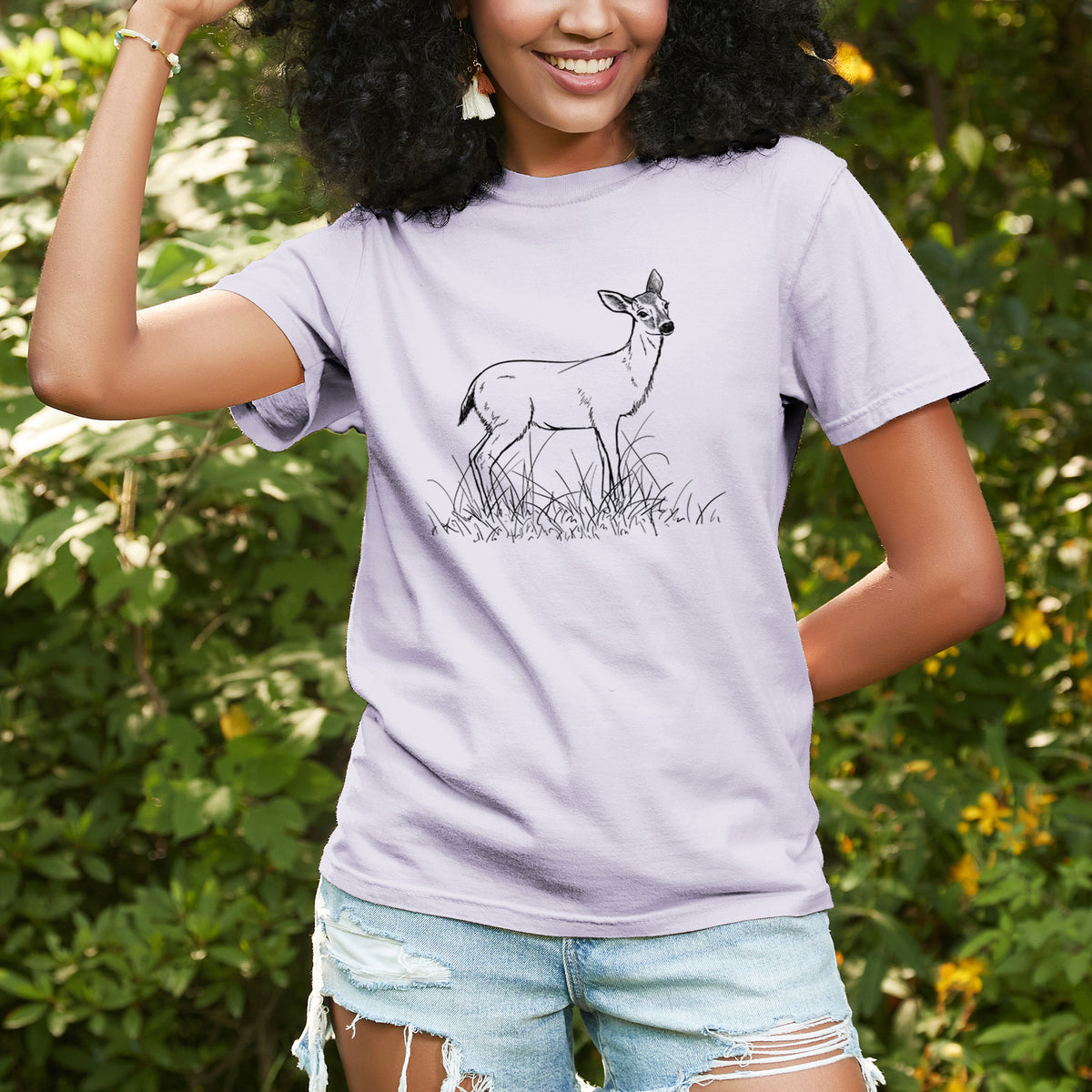 Woman wearing a Comfort Colors t-shirt printed with a black ink sketch of a black-tailed deer standing in tall grass, outdoors.
