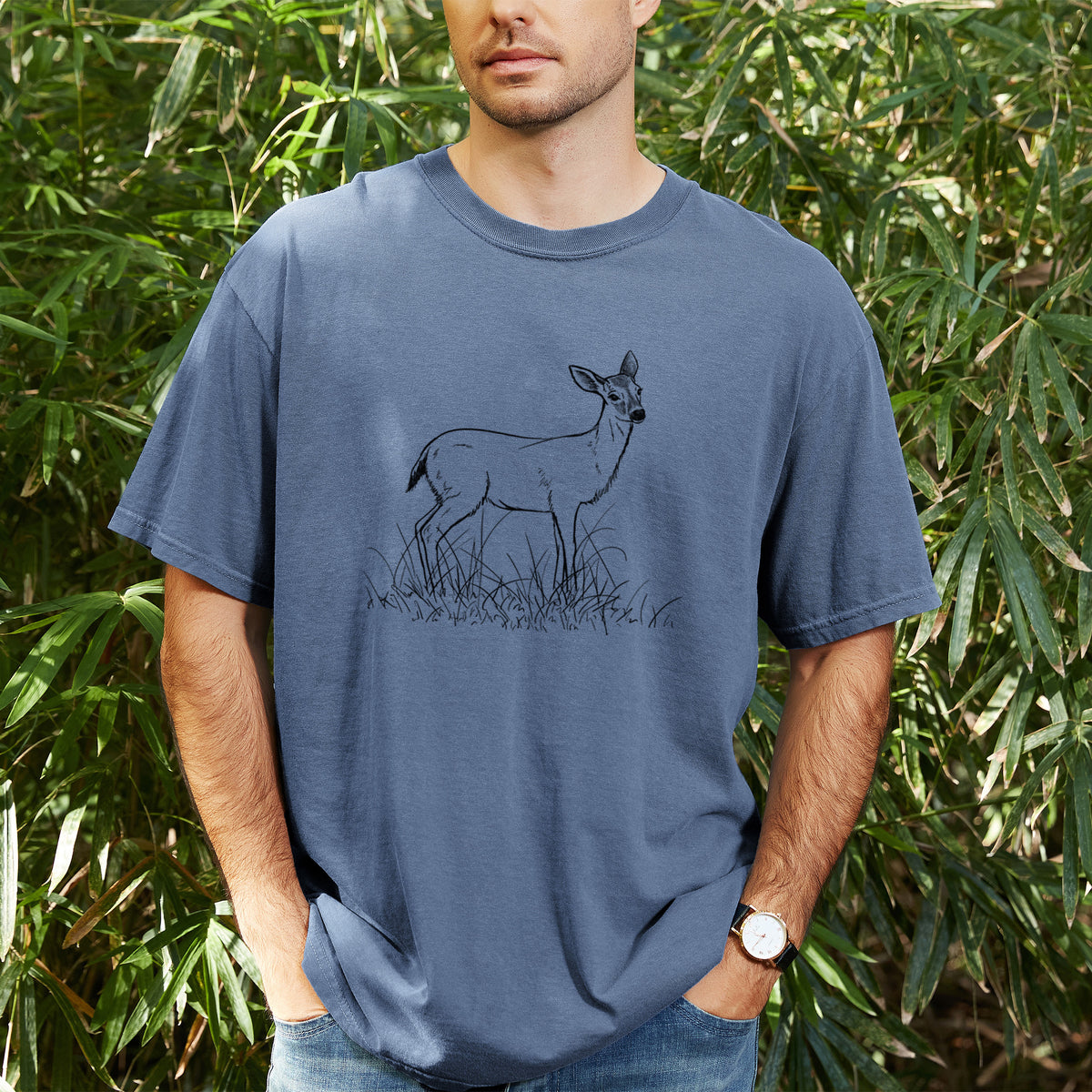 Man wearing a Comfort Colors t-shirt printed with a black ink sketch of a black-tailed deer standing in tall grass, outdoors.