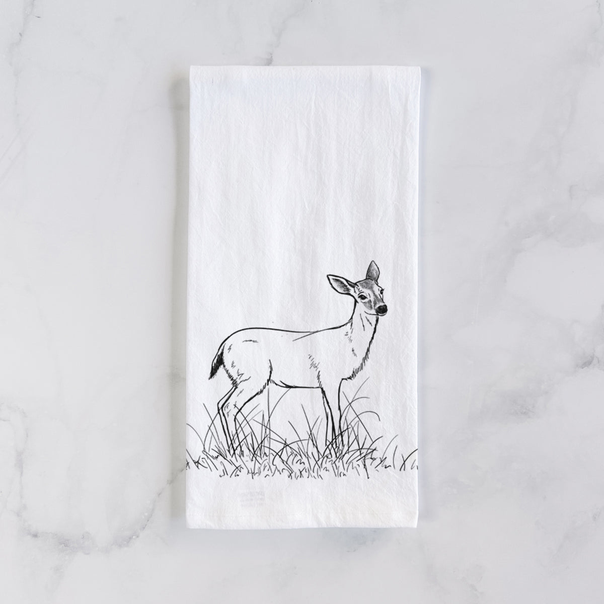 White cotton tea towel laid flat on marble, printed with a black ink sketch of a black-tailed deer standing in tall grass.