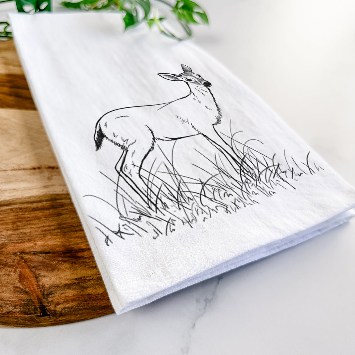Close-up of a folded white tea towel on a wooden board, showing a black ink sketch of a black-tailed deer standing in tall grass.