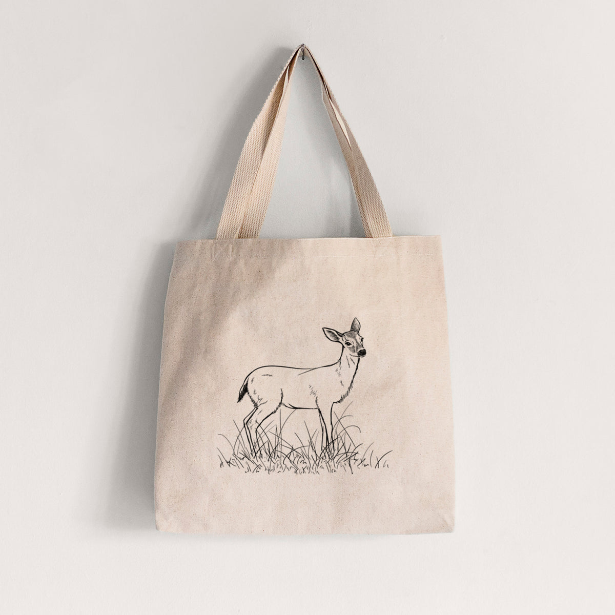 Natural canvas tote bag printed with a black ink sketch of a black-tailed deer standing in tall grass.