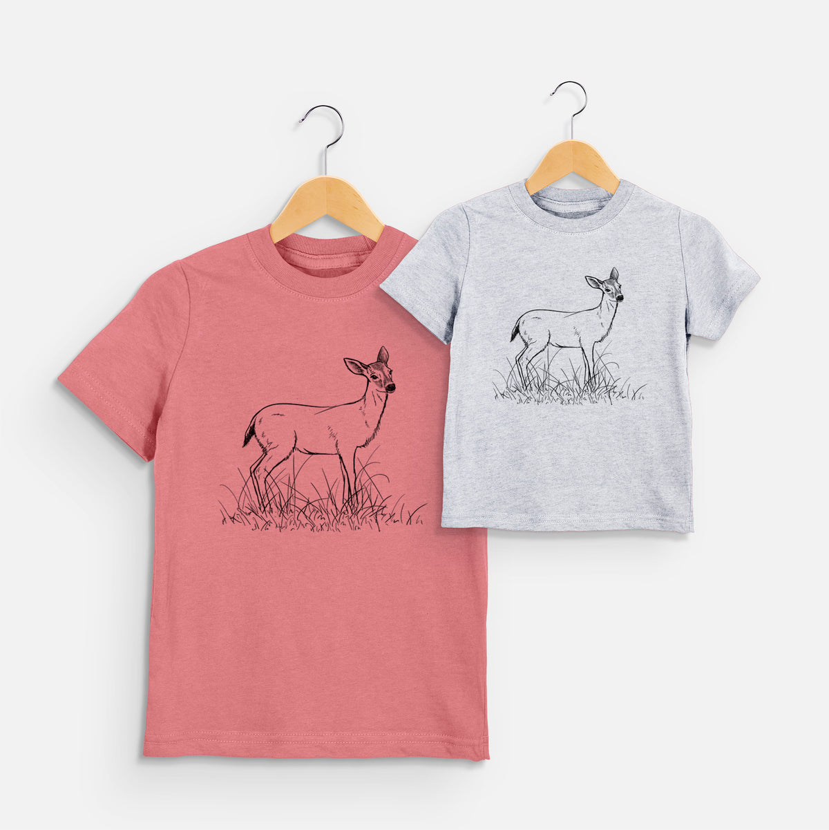 Two youth t-shirts in different colors on hangers, printed with a black ink sketch of a black-tailed deer standing in tall grass.