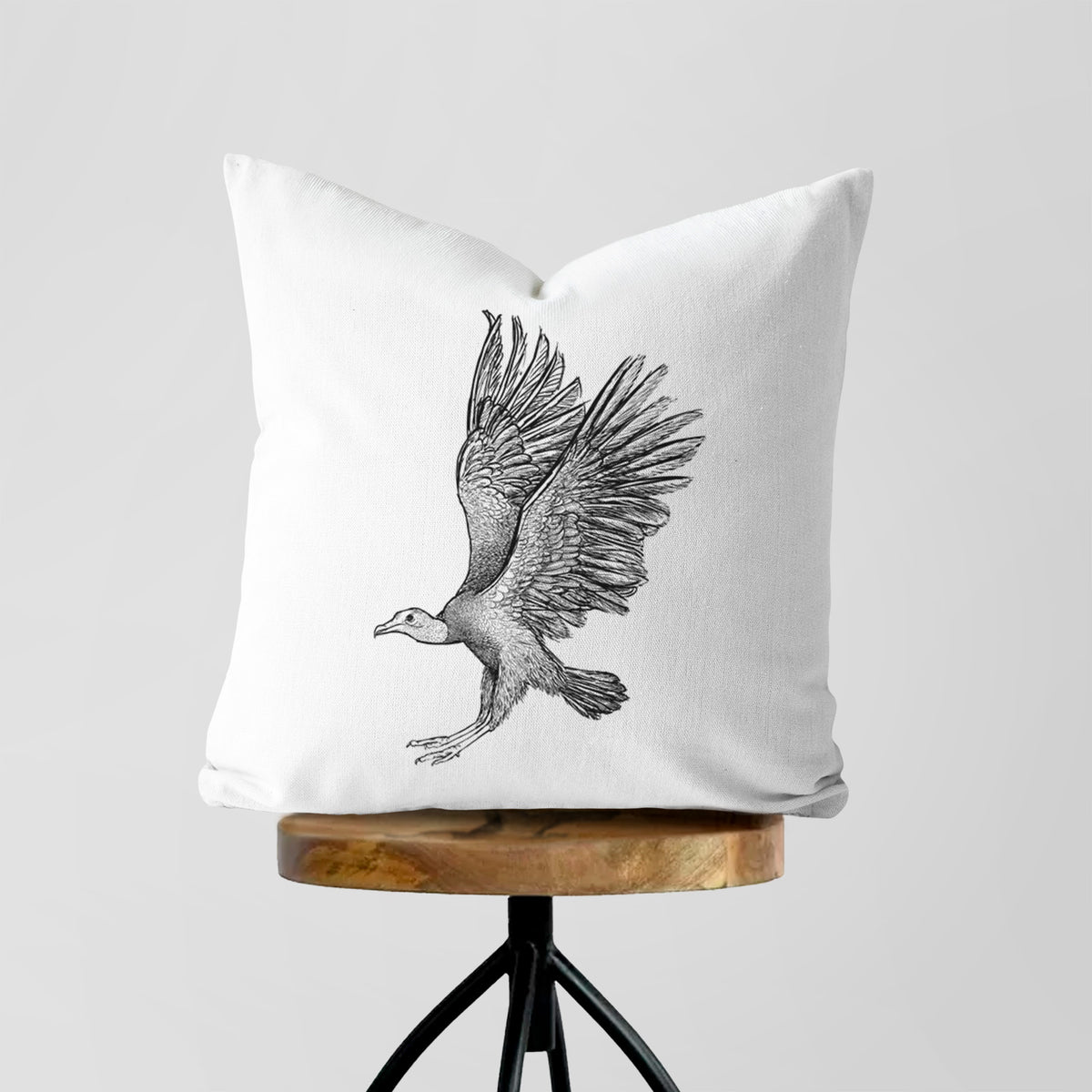 White pillow cover printed with a detailed black ink illustration of a black vulture landing with its wings spread, on a wooden stool.