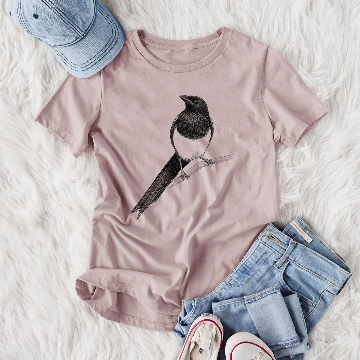 Women's relaxed fit cotton t-shirt printed with a detailed black ink sketch of a black-billed magpie perched on a branch with its long tail, styled flat on a white faux fur rug with jeans.
