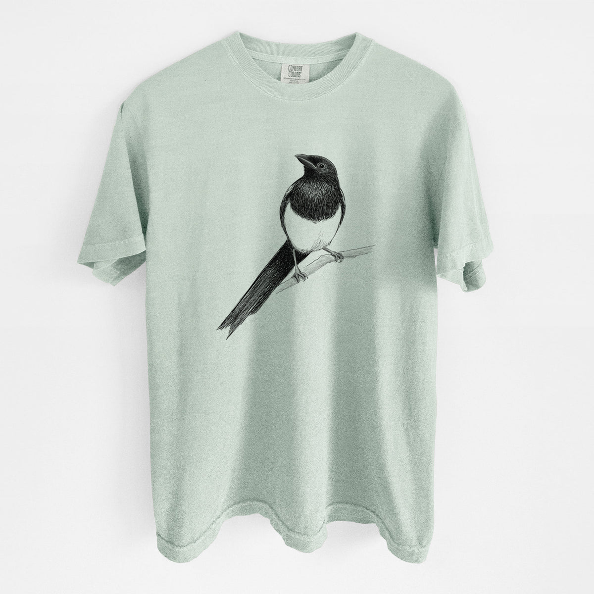 Bay green Comfort Colors t-shirt laid flat, printed with a detailed black ink sketch of a black-billed magpie perched on a branch with its long tail.