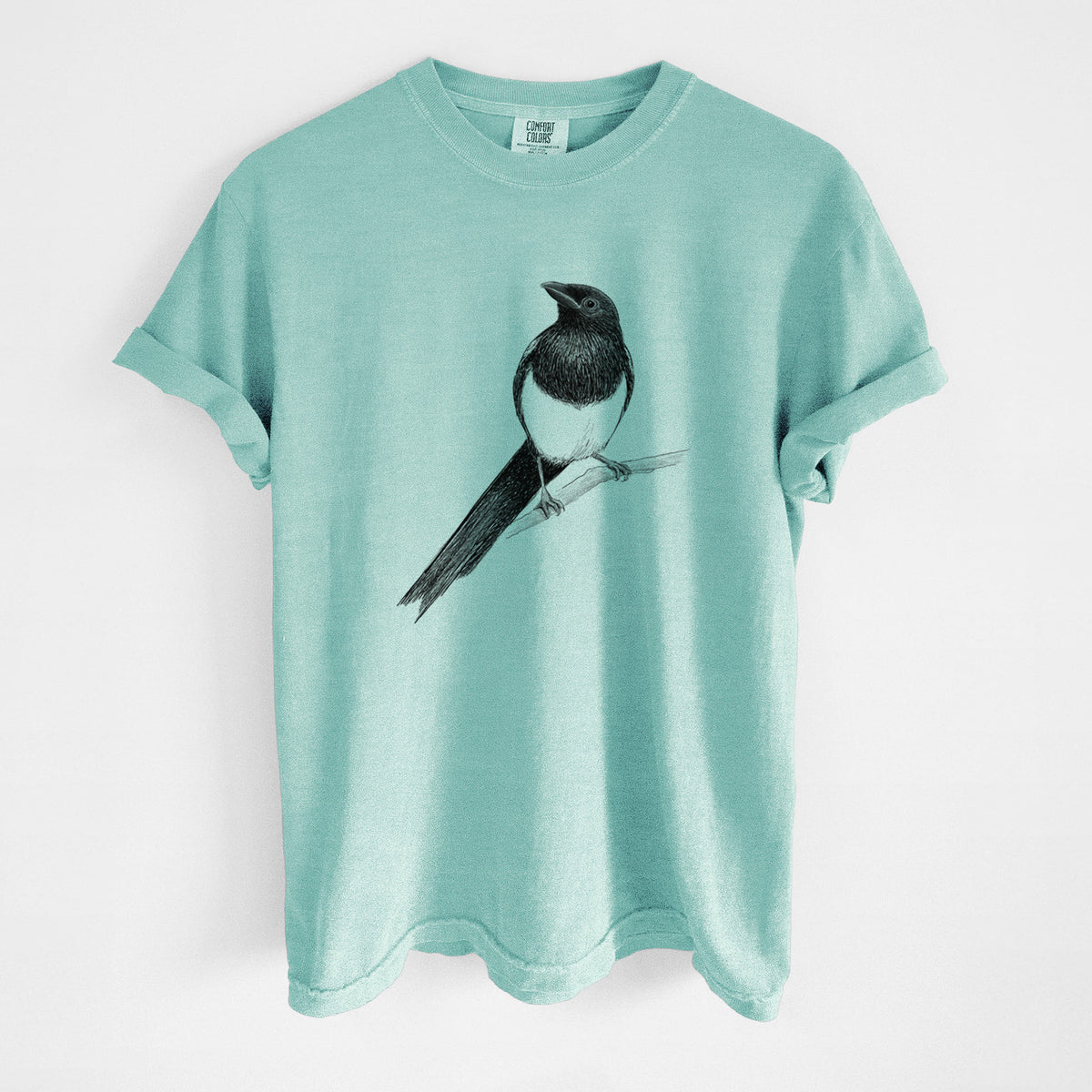 Chalky mint Comfort Colors t-shirt laid flat, printed with a detailed black ink sketch of a black-billed magpie perched on a branch with its long tail.