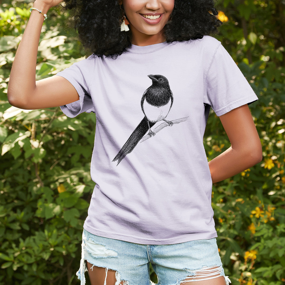Woman wearing a Comfort Colors t-shirt printed with a detailed black ink sketch of a black-billed magpie perched on a branch with its long tail, outdoors.