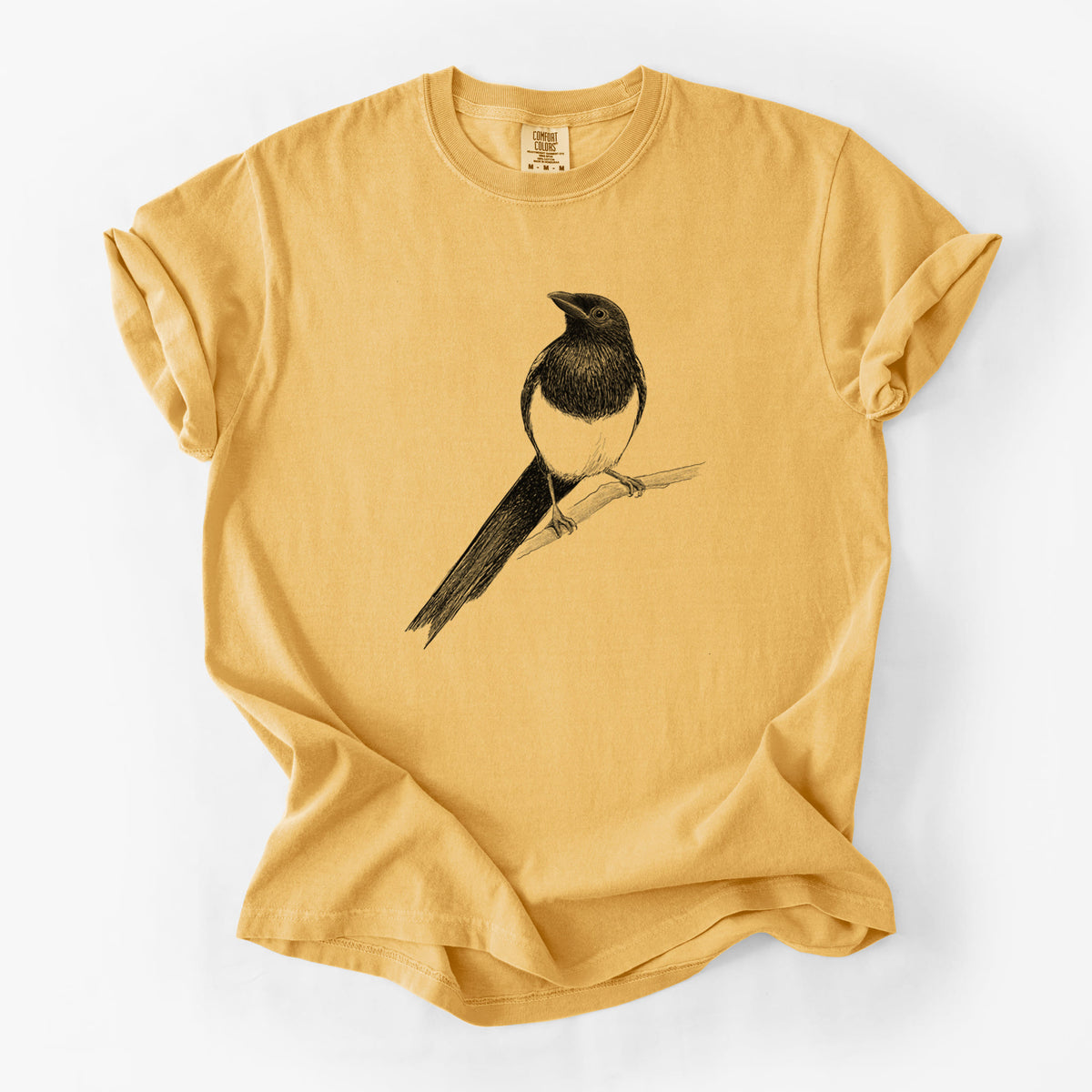 Golden wheat yellow Comfort Colors t-shirt laid flat, printed with a detailed black ink sketch of a black-billed magpie perched on a branch with its long tail.
