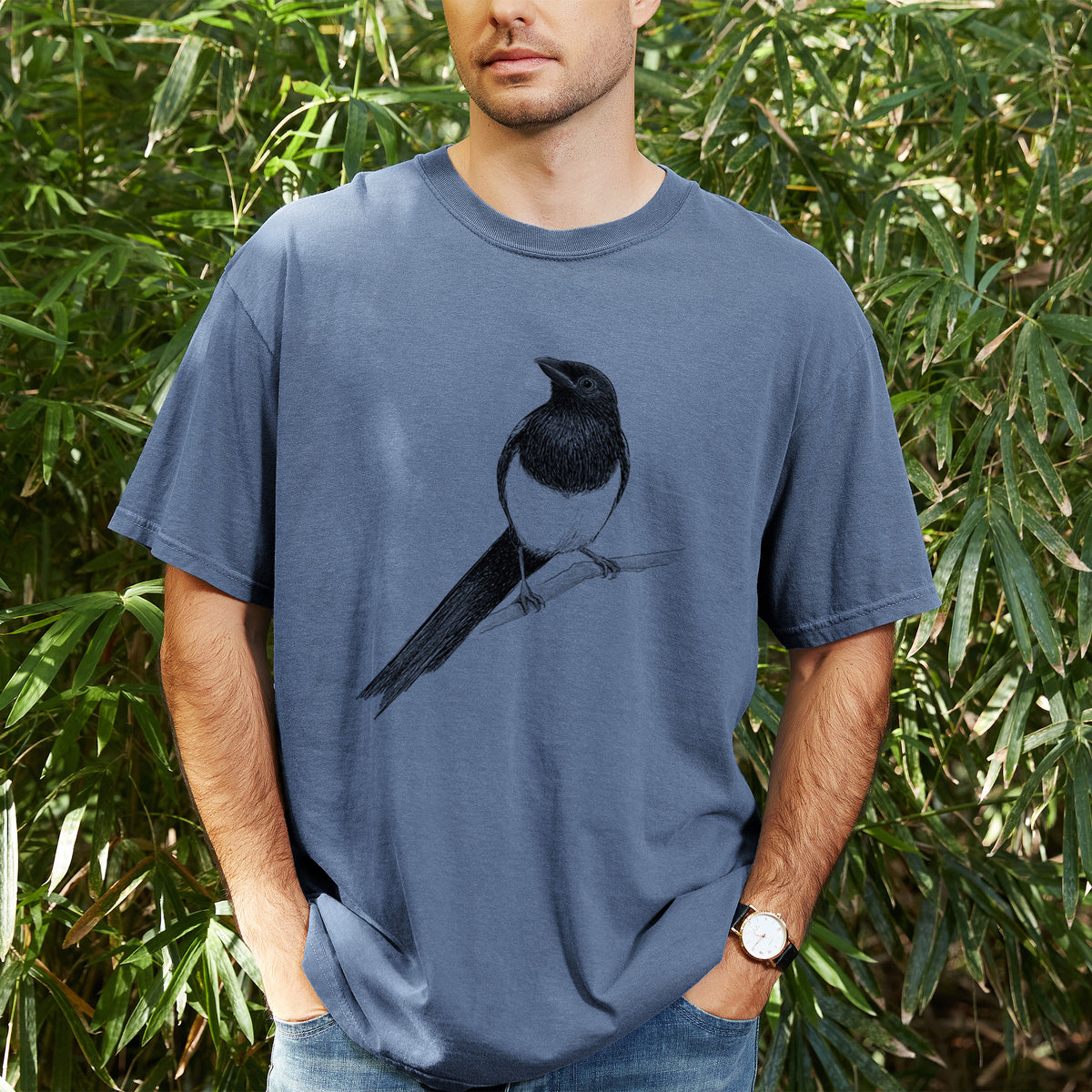 Man wearing a Comfort Colors t-shirt printed with a detailed black ink sketch of a black-billed magpie perched on a branch with its long tail, outdoors.