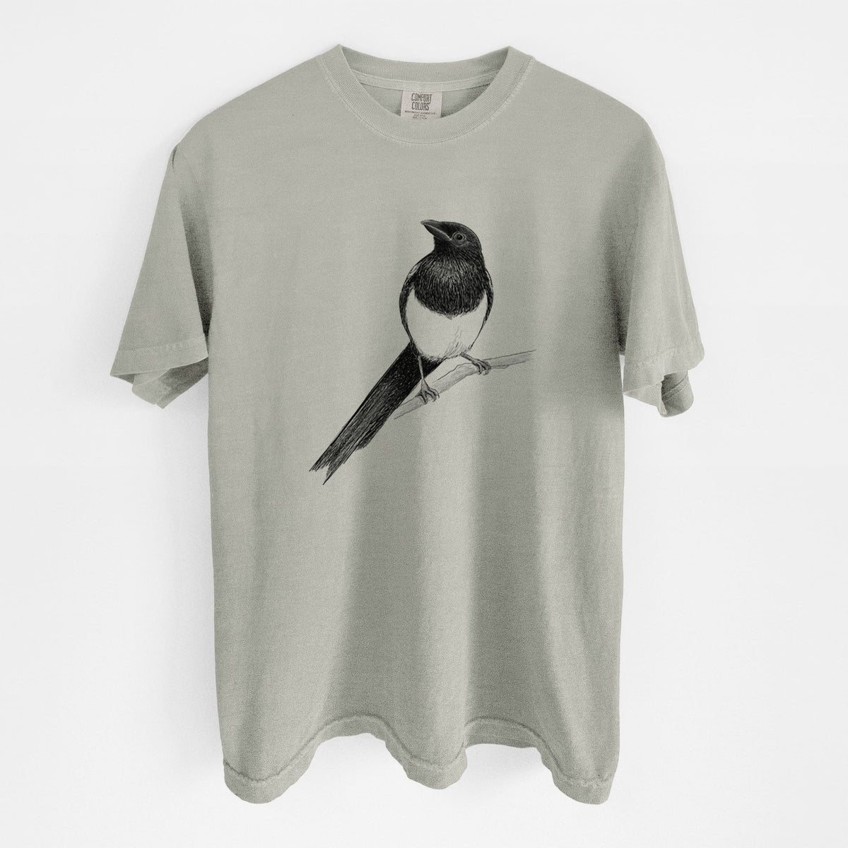 Sandstone beige Comfort Colors t-shirt laid flat, printed with a detailed black ink sketch of a black-billed magpie perched on a branch with its long tail.