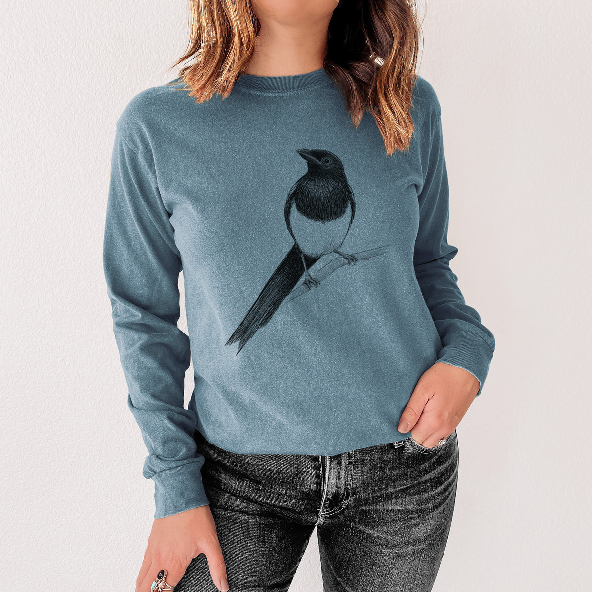 Steel blue Comfort Colors long sleeve shirt printed with a detailed black ink sketch of a black-billed magpie perched on a branch with its long tail, shown in a lifestyle photo.