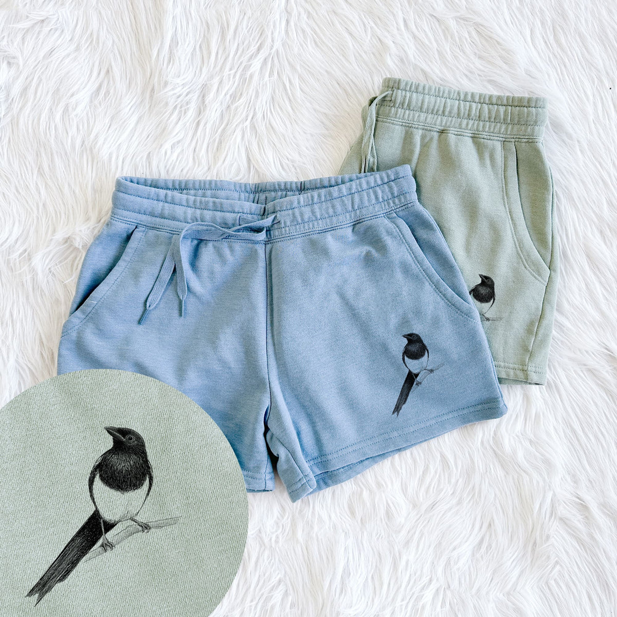 Two pairs of Cali Wave shorts in different colors printed with a detailed black ink sketch of a black-billed magpie perched on a branch with its long tail, with a close-up of the print.
