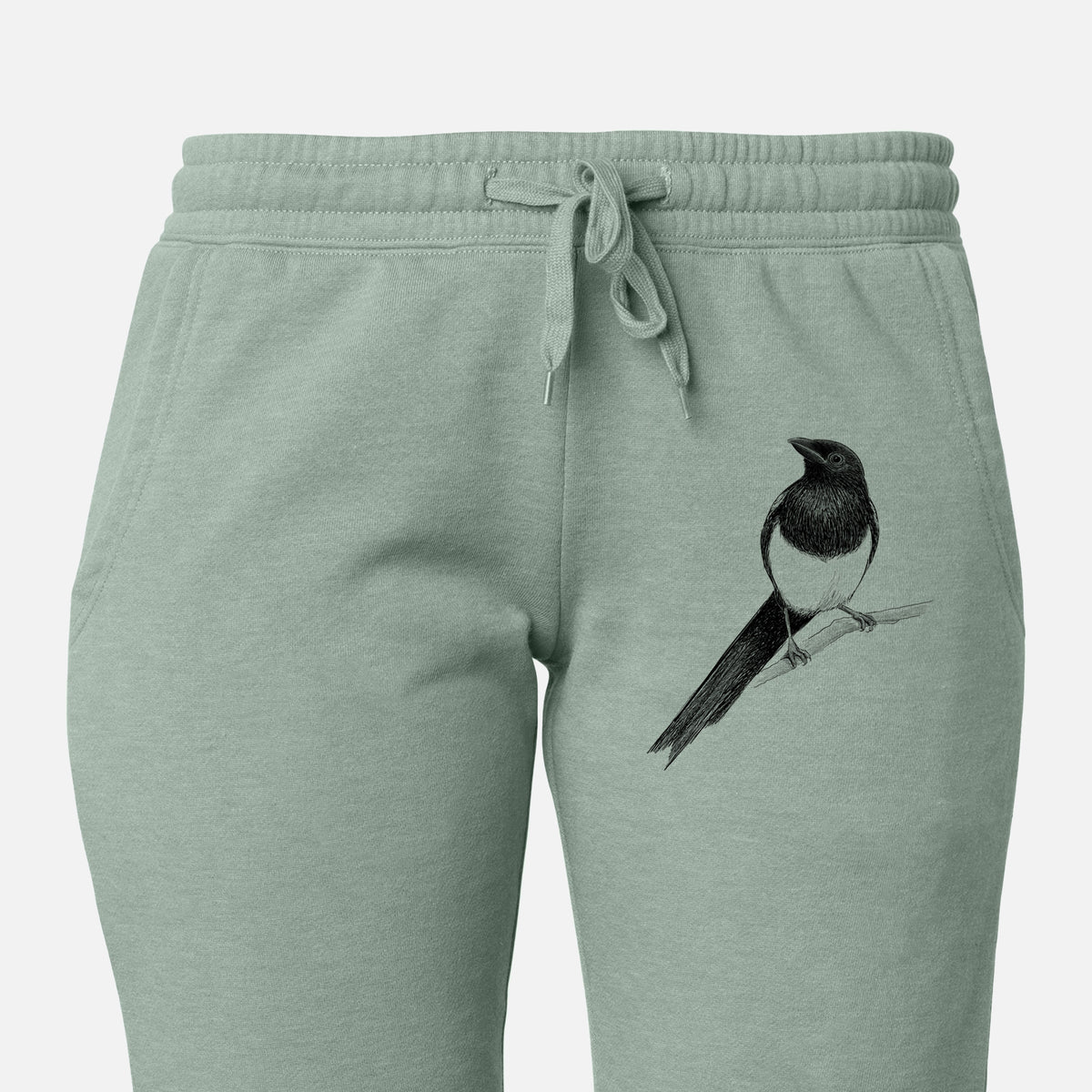 Front of sage green Cali Wave sweatpants, printed with a detailed black ink sketch of a black-billed magpie perched on a branch with its long tail on the leg.