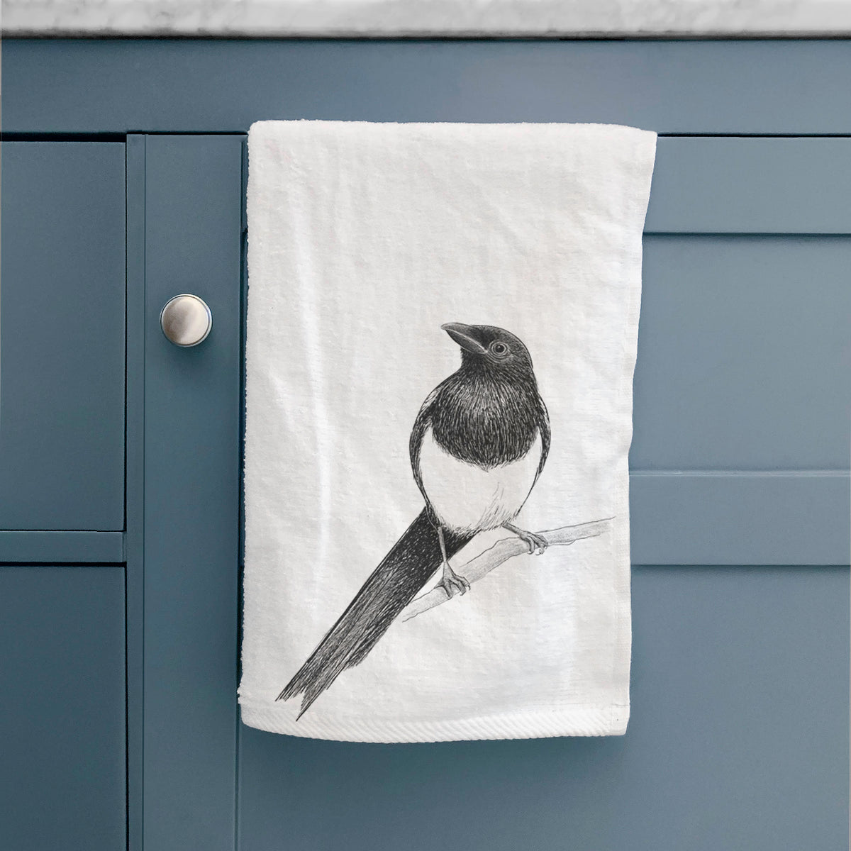 White hand towel printed with a detailed black ink sketch of a black-billed magpie perched on a branch with its long tail, hanging on a cabinet door.