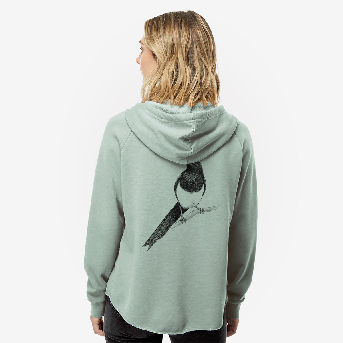 Woman wearing a sage green women's Cali Wave zip-up sweatshirt printed with a detailed black ink sketch of a black-billed magpie perched on a branch with its long tail, seen from behind.