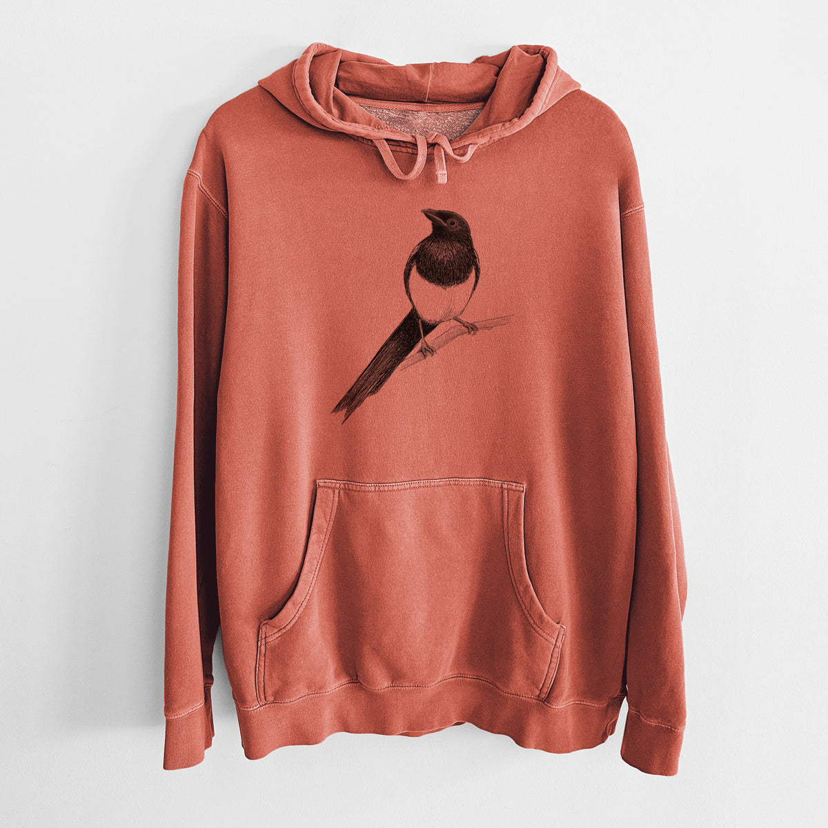 Amber orange pigment dyed hoodie laid flat, printed with a detailed black ink sketch of a black-billed magpie perched on a branch with its long tail.